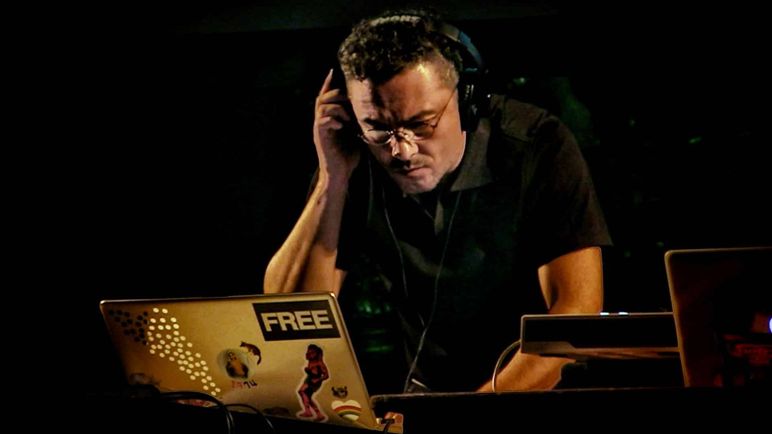 Carlos Adolfo Gutiérrez Vidal con audífonos y gafas, concentrado mientras manipula controles frente a una computadora portátil con calcomanías, en un ambiente oscuro de escenario.