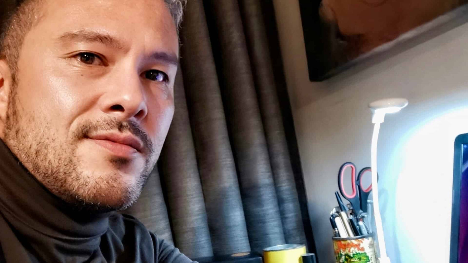 Primer plano de Carlos Adolfo Gutiérrez Vidal con suéter de cuello alto negro, sentado frente a un escritorio con lámpara encendida y utensilios de oficina al fondo.