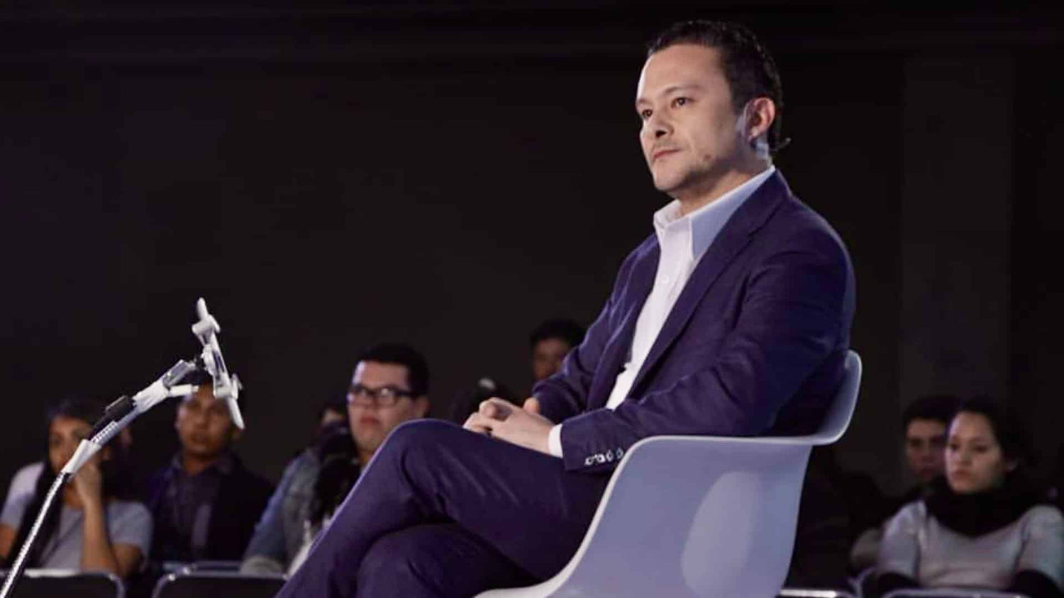 Carlos Adolfo Gutiérrez Vidal con traje azul marino y camisa blanca, sentado en un escenario frente a un micrófono, con público desenfocado al fondo.